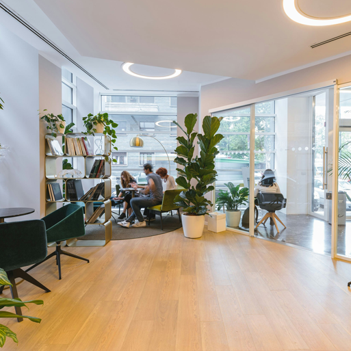 Bright modern office space with employees collaborating in a lounge area, large windows, indoor plants, and clean air-focused commercial workspace design.